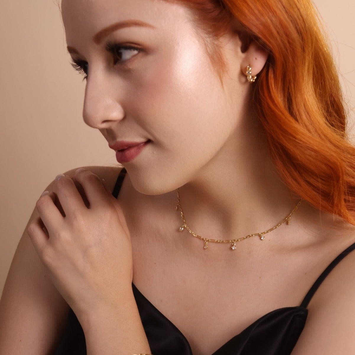 Woman with red hair wearing gold jewelry against a beige background